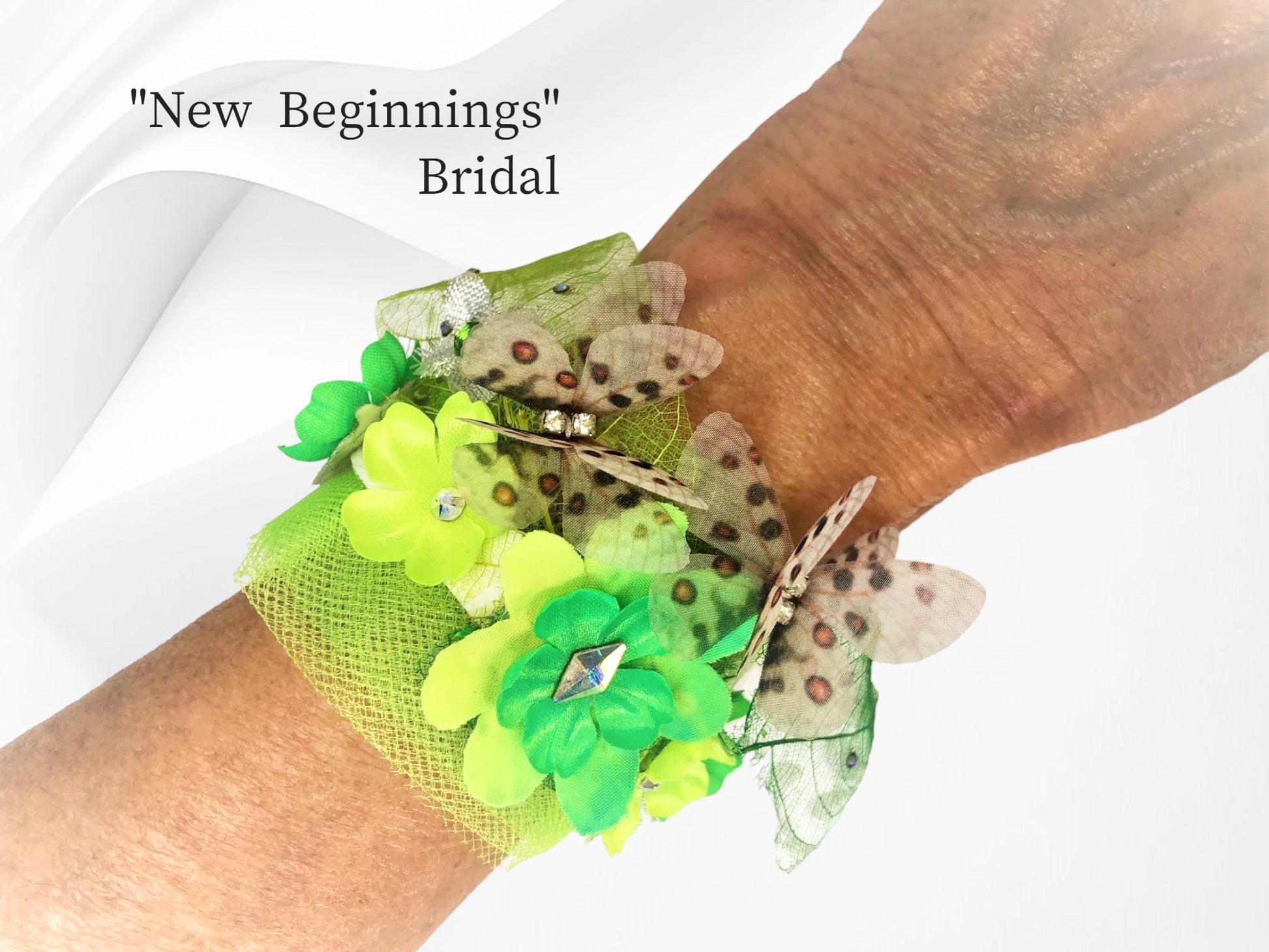 New Beginnings- Faux suede bracelet w/ magnetic closure. This bracelet has bright spring green silk flowers w/ matching Austrian Crystal centers & delicate tulle, bright green skeleton leaves w/ tiny crystal dew drops, metallic silver leaves, and 2 adorable silk butterflies w/ crystal centers. $42.99